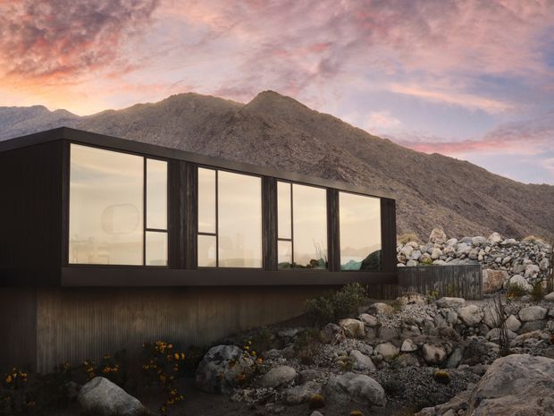 A modern dark-clad house set among a rocky mountain landscape. The low, rectilinear building features expansive floor-to-ceiling glazing, strong horizontal and vertical framing, and a partially elevated form above a rugged stone site. Warm sunset light reflects across the windows and surrounding mountain terrain.