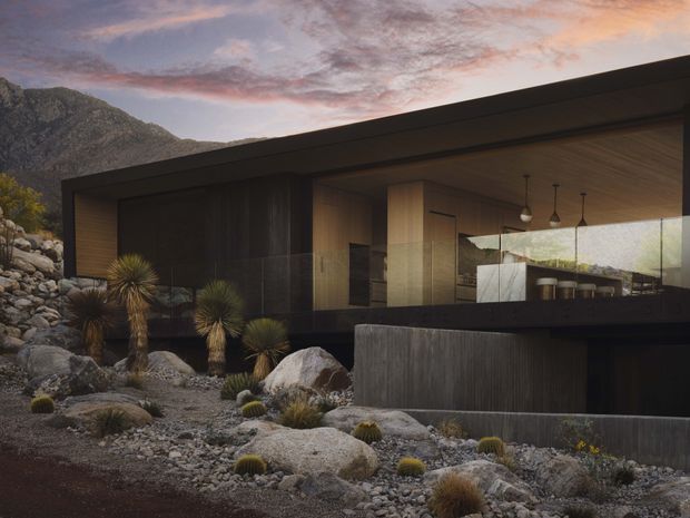 A contemporary, low-profile house integrated into a rugged mountain landscape. The dark horizontal facade combines broad glazing, timber-lined soffits and warm interior joinery, with a cantilevered roof and exposed concrete or dark cladding elements. Native drought-tolerant planting, boulders and gravel form the foreground landscape, while dramatic mountains and a sunset sky frame the building.