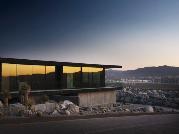 A low, modern waterfront building with a strong horizontal flat roof and extensive dark-framed glazing, set among rugged boulders and sparse landscaped planting. The reflective windows capture warm sunset light, while a bay, illuminated shoreline, and mountainous backdrop are visible beyond.