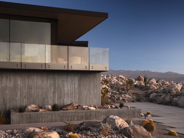 Exterior view of a modern, low-profile building set within a rocky desert landscape. The structure features a strong cantilevered flat roof, exposed textured concrete walls, expansive glazing, and a transparent glass balustrade on an upper terrace. Large boulders, desert plants, paved outdoor paths, and distant rugged mountains surround the building.
