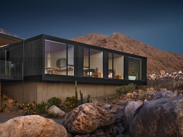 A modern single-storey residence set into a rocky desert mountain landscape. The home features a dark vertically ribbed exterior, a low horizontal form, extensive floor-to-ceiling glazing, and a projecting roof overhang. Warmly lit interiors are visible through the windows, while large boulders, cacti, and sparse planting surround the building.