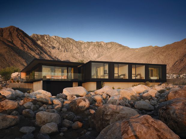 A low, modern single-storey residence set among large rounded desert rocks, with dark horizontal framing, extensive floor-to-ceiling glazing, warm timber-clad lower walls, and dramatic rugged mountain terrain behind it.