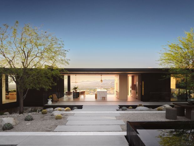 A low-slung modern single-storey residence viewed from a landscaped courtyard. The dark flat-roofed facade opens through extensive glazing and sliding doors to a bright interior, with a long central axis of pale concrete stepping slabs, gravel landscaping, sculptural desert plants and a reflecting pool in the foreground. Mountains are visible beyond the house.