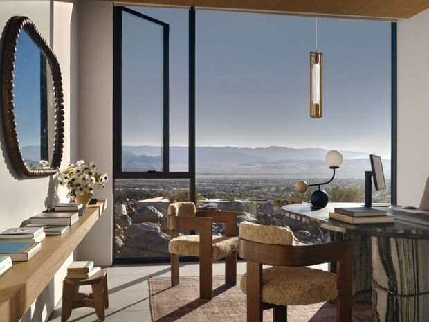 A refined modern interior with floor-to-ceiling corner glazing overlooking a broad mountain and valley landscape. The room features a timber ceiling, warm wood shelving and furniture, sculptural boucle-upholstered dining chairs, a stone or marble-clad console, a decorative pendant light, and an ornate dark-framed mirror. The space appears to combine a dining or sitting area with a compact workspace, creating an airy, warm, sophisticated atmosphere.
