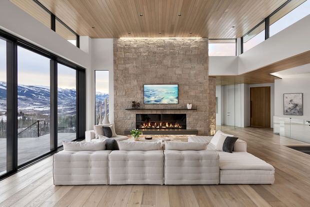 Spacious contemporary living room with a large modular cream sectional sofa facing a linear fireplace set into a full-height natural stone feature wall. The room has extensive floor-to-ceiling glazing overlooking a snowy mountainous landscape, a warm timber-lined ceiling and timber flooring, recessed downlights, and an open-plan connection to a hallway and adjacent spaces. The design is refined, warm, and strongly connected to the alpine setting.
