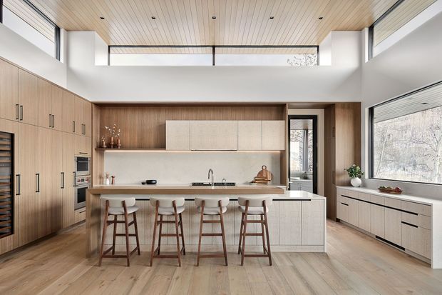 Bright, spacious contemporary kitchen with a large central island and five timber stools. Full-height warm timber cabinetry lines the left wall, while pale textured cabinetry and drawers extend beneath a broad window on the right. The island includes a light stone benchtop and integrated cabinetry, with a pale stone or tiled splashback behind the cooktop and sink. A slatted timber ceiling, clerestory glazing, broad windows, and light timber flooring create an airy, warm interior.
