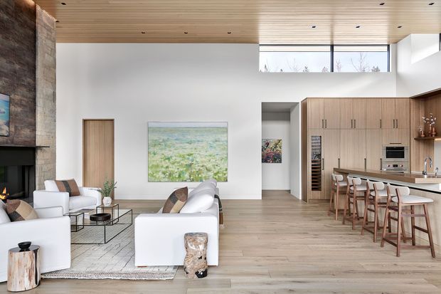Open-plan contemporary living, dining, and kitchen space with a double-height ceiling, warm timber ceiling lining, pale timber flooring, extensive natural light, and large fixed timber cabinetry. A white sectional sofa and armchairs define the living area, while a long kitchen island with timber stools occupies the right side. A dark stone or concrete fireplace wall anchors the left side, and high clerestory glazing brightens the room.