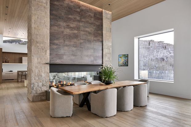 Open-plan dining area with a long live-edge timber dining table and upholstered chairs positioned in front of a dramatic stone-clad fireplace. The room features a timber-lined ceiling, pale timber flooring, broad grey plaster walls, large windows, and an open view toward the kitchen. A linear fire is visible behind the dining table, with a large textured stone feature wall extending to the ceiling. The overall design is warm, contemporary, and refined.