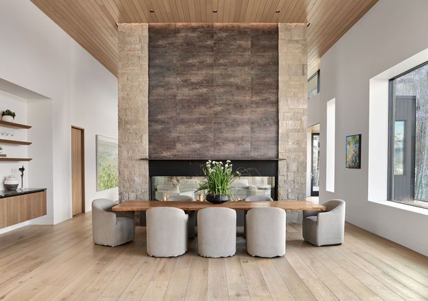 Bright, spacious open-plan dining area centred on a long natural timber dining table with upholstered curved chairs. A tall stone-clad fireplace wall with dark textured feature cladding forms the focal point, while a timber-lined ceiling, pale timber flooring, white walls, large windows and built-in shelving create a warm contemporary interior.