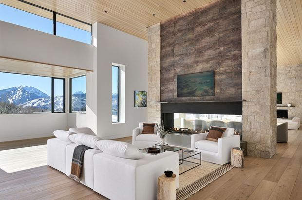 A spacious, light-filled contemporary living room with a double-height ceiling and expansive windows framing snow-covered mountains. The room features a large white sectional sofa, white lounge chairs, a glass coffee table, a linear fireplace set within a tall rugged stone feature wall, and warm timber flooring and ceiling lining. An open mezzanine or upper-level void is visible above the living area.
