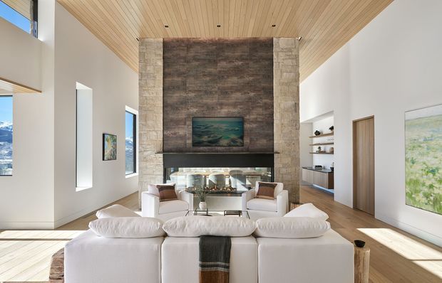 Bright, spacious contemporary living room viewed from behind a large white sectional sofa. The room features a full-height stone fireplace wall with a dark timber or stone feature panel, built-in shelving, large windows, white walls, a warm timber-lined ceiling, and light timber flooring. Two armchairs and a glass coffee table face the fireplace, with artwork and mountain scenery visible through the windows.