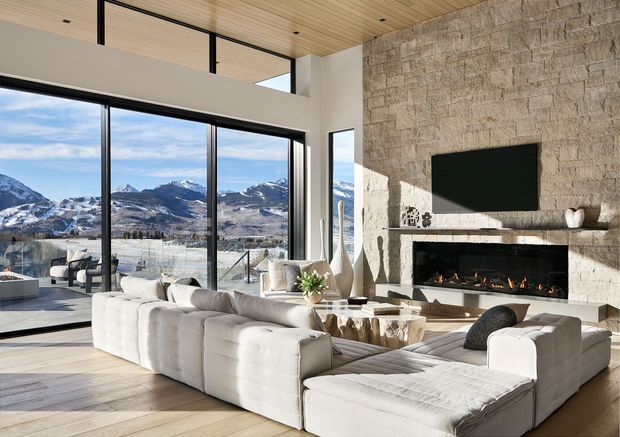 A bright, spacious contemporary living room with floor-to-ceiling glazing overlooking snow-covered mountains. The room features a large white modular sectional sofa, pale timber flooring and ceiling, black-framed glazing, and a substantial textured natural-stone fireplace wall with a built-in linear fire and wall-mounted television. A mezzanine-like upper opening and high glazing contribute to the expansive volume.