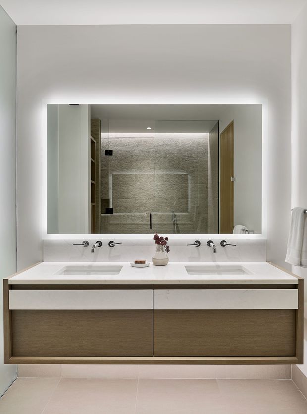 Contemporary bathroom vanity with a wide backlit mirror, double undermount basins, wall-mounted chrome tapware, and a floating timber-look drawer cabinet. A frameless glass shower with textured pale tile walls is reflected in the mirror. The room uses clean horizontal lines, soft neutral finishes, and concealed lighting.