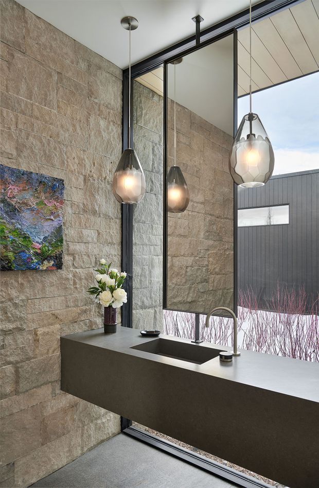 A contemporary kitchen or kitchenette with a long dark stone island and integrated sink, set against a full-height textured natural-stone wall. Black-framed floor-to-ceiling glazing and a large mirror reflect the stone surfaces and pendant lights. Three sculptural smoked-glass pendant lights hang above the island, creating a refined, dramatic atmosphere. An exterior courtyard or adjacent building is visible through the glazing.