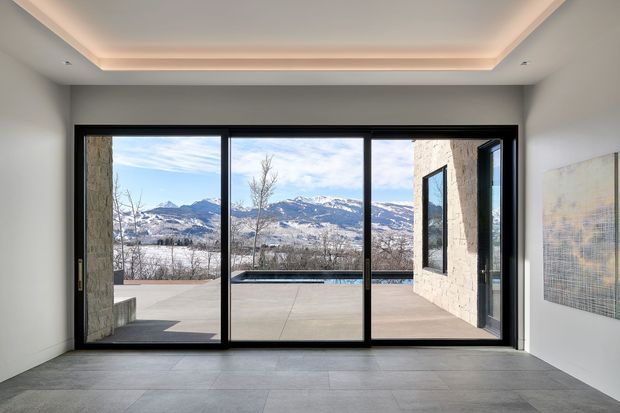 A minimalist contemporary interior looking through expansive black-framed floor-to-ceiling glazing toward a broad outdoor terrace and mountain landscape. The room has large-format grey tiled flooring, smooth light walls, a recessed tray ceiling with concealed warm cove lighting, and a framed artwork on the right wall. The glazing opens onto a paved terrace with a narrow linear pool or water feature visible beyond the threshold.