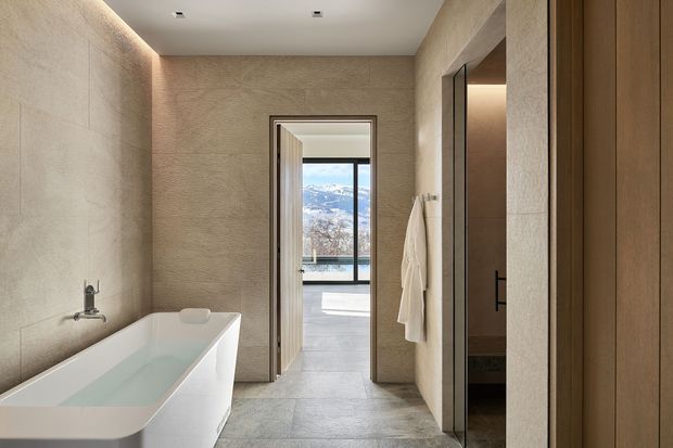 A refined contemporary bathroom with a freestanding white bathtub, large-format pale stone-look floor tiles, and textured beige wall finishes. A wall-mounted tap is positioned above the bath. The room opens directly toward a bright glazed space with a mountain view, while a partial vanity area is visible through the opening on the right. Recessed ceiling lights and concealed linear lighting create a warm, calm atmosphere, with timber-toned joinery or door panels adding contrast.