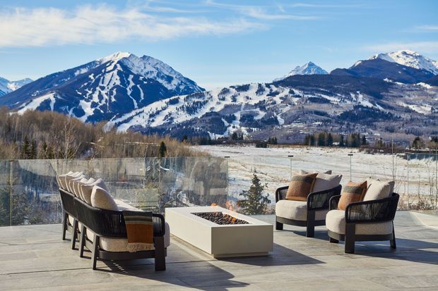 An outdoor terrace or deck furnished with lounge chairs and a long dining-style seating arrangement around a rectangular fire table. A transparent glass balustrade frames expansive snow-covered mountain views, with a pale paved surface and sparse landscaping in the foreground. The scene is bright, contemporary, and designed for outdoor entertaining in an alpine setting.