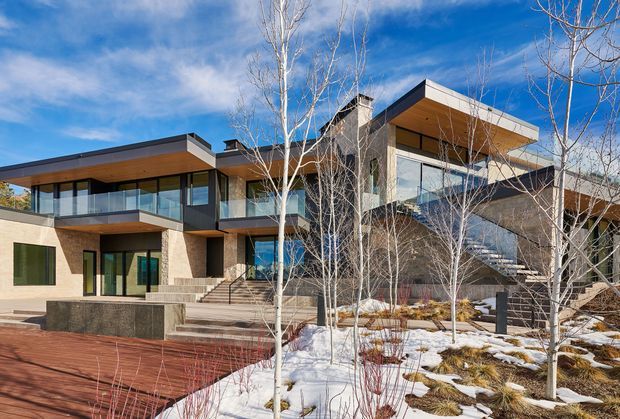 Exterior view of a large contemporary multi-level residence with flat roof planes, extensive floor-to-ceiling glazing, cantilevered upper volumes, balconies with glass balustrades, and warm timber soffits. The house combines pale masonry and dark structural or cladding elements, with broad steps leading to a landscaped terrace and a snow-covered garden with slender deciduous trees.