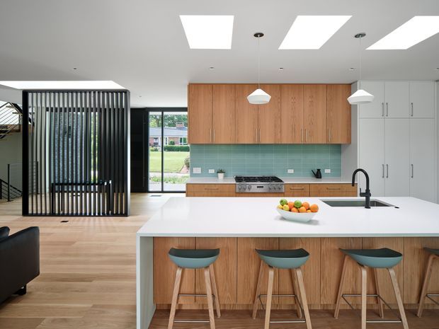 Contemporary open-plan kitchen with a large white island and seating for three, warm timber cabinetry, a pale green tiled splashback, white full-height storage, and light timber flooring. Black-framed glazing and a dramatic vertical slatted partition define the adjoining living area, while skylights and pendant lights provide bright illumination.