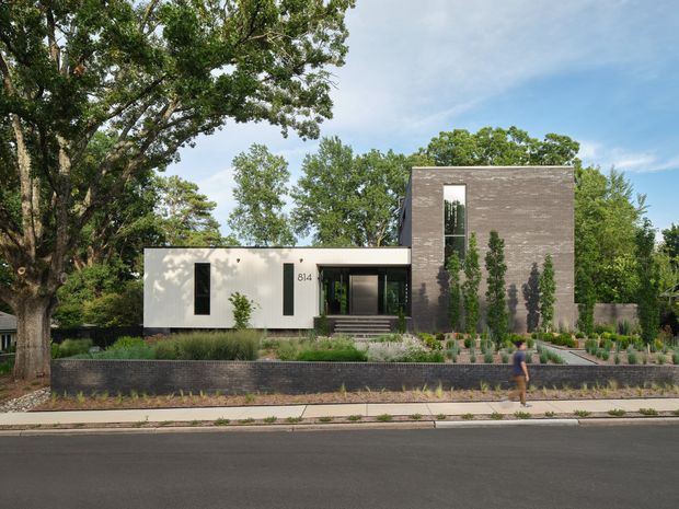 Street-facing view of a contemporary two-part residence set within a landscaped suburban site. The home combines a low, flat-roofed white volume with a taller dark masonry-clad rectangular volume, narrow vertical windows, and a recessed entry approached by steps. A low dark retaining wall, layered planting, mature trees, sidewalk, and street are visible in front.