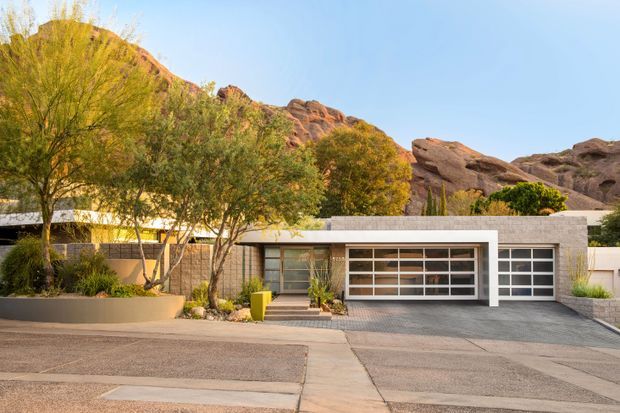 Street-facing exterior view of a low, single-storey residence with a broad flat roof, pale masonry walls, attached double garage, and large horizontal garage glazing. The landscaped frontage includes mature desert trees and raised planters, while dramatic rocky hills rise immediately behind the house.