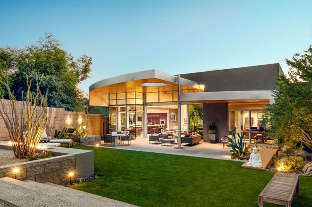 A contemporary single-storey residence viewed from a landscaped backyard. The home features broad floor-to-ceiling glazing, a flat roof with strong horizontal planes, warm timber-toned soffits, and an open indoor-outdoor living area opening onto a paved terrace. The foreground includes a manicured lawn, raised masonry planters, garden lighting, sculptural planting, and a timber bench. The image has a warm, polished dusk atmosphere.
