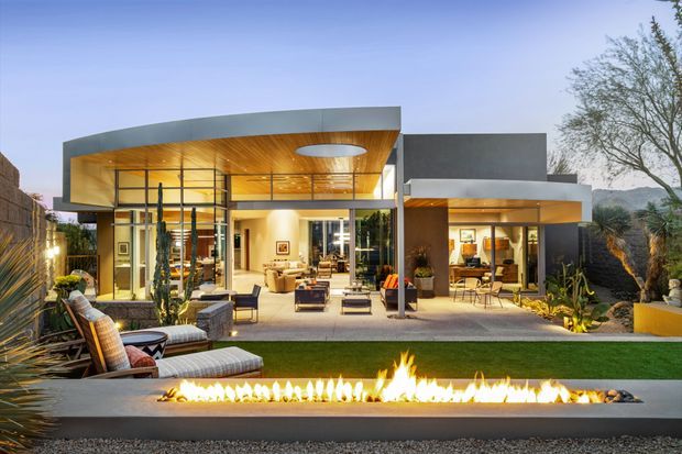 Exterior view of a contemporary single-level residence at dusk, featuring broad floor-to-ceiling glazing, a projecting flat roof with a warm timber-lined soffit, dark rendered volumes, and a landscaped outdoor entertaining area. The foreground includes a long linear outdoor fire feature, lawn, lounge seating, planting, and a paved terrace. Interior living and dining spaces are visible through the glazing, with hills and trees in the distant background.