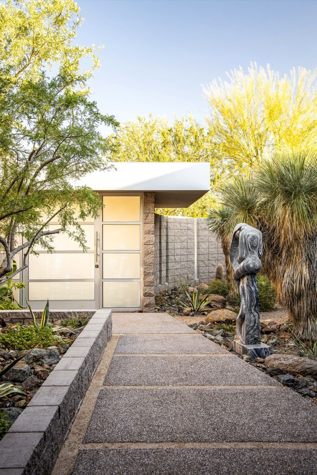A landscaped exterior entry approach leads to a compact modern building with a crisp white low-slope roof canopy, translucent glazed entry doors, and a textured stone-clad wall. The pathway is formed from exposed aggregate or textured concrete with darker dividing bands and bordered by low masonry edging. Drought-tolerant planting, ornamental grasses, rocks, and a sculptural figure create a natural desert-style garden setting.