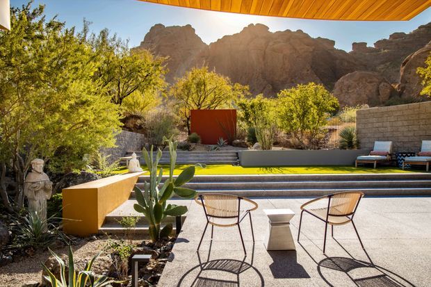 A sunlit outdoor terrace and pool area framed by a timber-lined roof overhang, with sculptural lounge chairs, a low angular bench, concrete paving, planting beds and desert landscaping. A long narrow pool sits beyond the terrace, backed by rendered and block walls, mature trees and dramatic rocky mountain formations. The setting has a warm, resort-like desert character with strong indoor-outdoor connection.