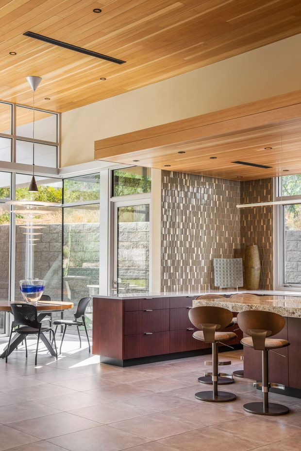 A bright, open-plan kitchen and dining area with extensive floor-to-ceiling glazing, warm timber-lined ceilings, dark wood cabinetry, and a patterned textured feature wall. A stone island with rounded brown bar stools dominates the foreground, while a small dining table and chairs sit beside the windows. The space has a refined mid-century-inspired feel with strong natural light and garden views.