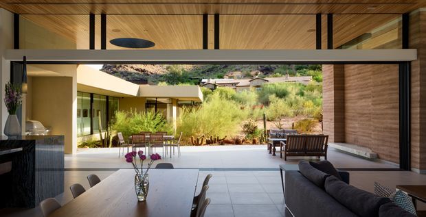Open-plan contemporary living and dining space extending onto a covered outdoor terrace or courtyard. The room features extensive sliding glass openings, a timber-lined ceiling, dark vertical structural or trim elements, pale large-format floor tiles, a timber dining table, and a dark sectional sofa. A landscaped dry-climate garden and neighbouring hillside homes are visible beyond the open glazing.