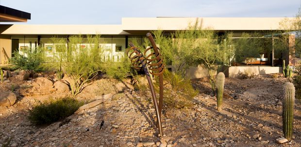 Exterior view of a low, single-storey modern residence set within a dry desert landscape. The building has a long, flat roofline, pale rendered walls, extensive dark-framed glazing, and sheltered outdoor areas. The foreground features gravel, exposed rock, drought-tolerant shrubs, tall cacti, and a sculptural metal garden artwork.