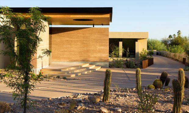 A contemporary single-storey residence in a dry, desert-like landscape, featuring flat rooflines, warm timber-clad walls, pale rendered walls, broad outdoor terraces and steps, and a sheltered entry courtyard. Native planting and tall columnar cacti define the foreground landscape.