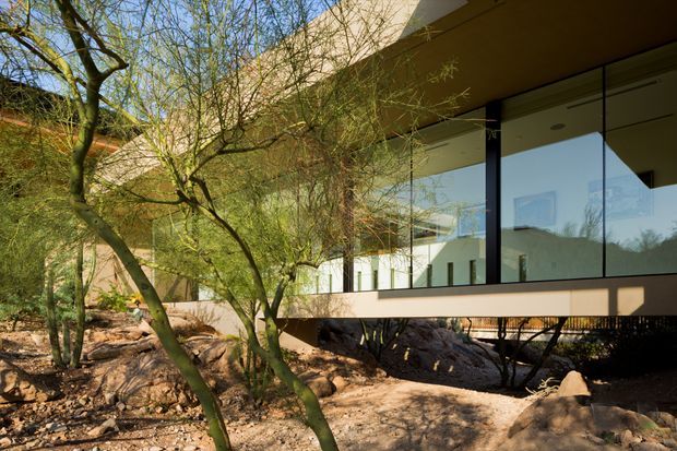 Exterior architectural photograph of a modern, elevated building set within a rocky, tree-filled landscape. The structure features extensive floor-to-ceiling glazing, slim dark window frames, a projecting flat roof, and a raised horizontal volume supported above the natural terrain.