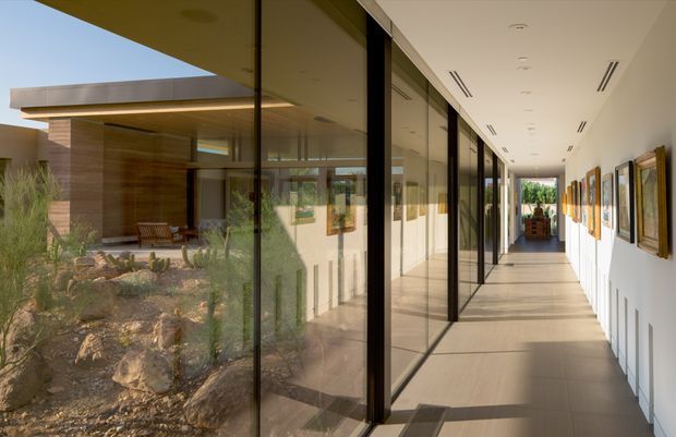 A long, bright contemporary gallery-like hallway with floor-to-ceiling glazing on one side, offering views into a landscaped courtyard and an adjacent timber-clad pavilion. The corridor has pale smooth flooring, white walls, slim black window and door frames, recessed linear ceiling vents and lighting, and framed artwork displayed along the right wall. Strong sunlight creates a clean, airy atmosphere and emphasizes the indoor-outdoor connection.