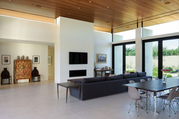 A bright, spacious open-plan living and dining area with a contemporary architectural interior. The room features a large dark sectional sofa facing a wall-mounted television and linear fireplace, a dining table with modern molded chairs, full-height black-framed glazing, white walls, pale tiled flooring, and a warm timber-lined ceiling with recessed lighting. Decorative furniture and artwork add traditional accents at the rear of the room.