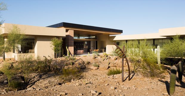 A low-slung contemporary single-storey residence set in a dry desert landscape. The building has flat roof planes, warm beige rendered walls, dark horizontal roof fascia, extensive glazing, and a sheltered central outdoor living area. Native desert planting, gravel, rocks, and sculptural metalwork surround the home.