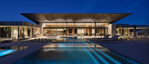 A dramatic twilight view of a low-slung contemporary residence arranged around a long rectangular swimming pool. The home features extensive floor-to-ceiling glazing, a broad projecting flat roof with a warm timber-lined soffit, dark exterior walls and paving, and a central open-plan living area visibly connected to the pool terrace. Landscape planting and lounge furniture frame the pool, while the deep blue sky and illuminated interiors create a luxurious, resort-like atmosphere.