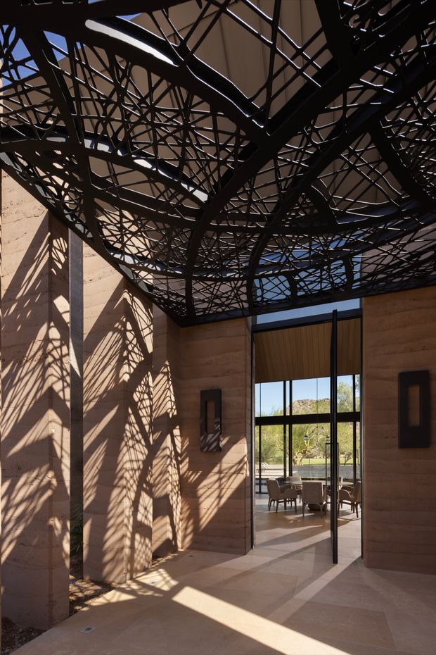 A dramatic architectural interior entry or foyer with tall sandstone-coloured masonry walls and a monumental black steel canopy overhead. The canopy has an intricate branching lattice that casts strong organic shadows across the walls and stone floor. A large glazed opening leads to a furnished room beyond, with views of a dry, rocky landscape. The space is sunlit, sculptural, and highly geometric, with a strong contrast between warm stone and dark metal.