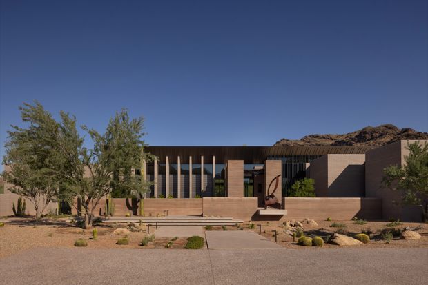 A low-slung contemporary desert residence viewed from the front, with a warm brown masonry and timber-toned facade, deep roof overhangs, vertical screened glazing, broad steps, and a sparsely planted xeriscape garden. Rocky terrain rises behind the building under a clear blue sky.