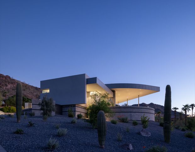 Exterior view of a contemporary single-storey residence set in a dry desert landscape at dusk. The house features strong rectilinear volumes, a broad curved roof canopy, extensive warm recessed lighting, and curved masonry walls. Gravel landscaping with columnar cacti, palms, and sparse drought-tolerant planting surrounds the home, with rocky hills visible behind.