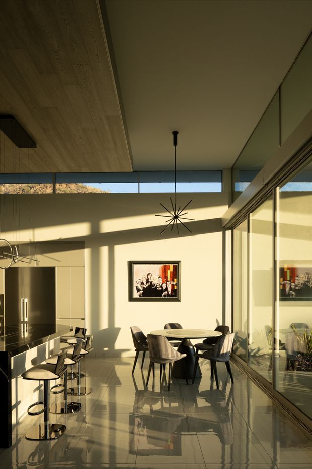 A sunlit open-plan kitchen and dining area with a high ceiling, clerestory glazing, and extensive glass walls. The kitchen has a dark island with a reflective countertop and a row of modern bar stools, while a round dining table with contemporary chairs sits centrally. Warm sunlight creates strong shadows across the pale walls and glossy tiled floor. A sculptural starburst pendant hangs above the dining table, with artwork and reflected planting visible through the glazing.