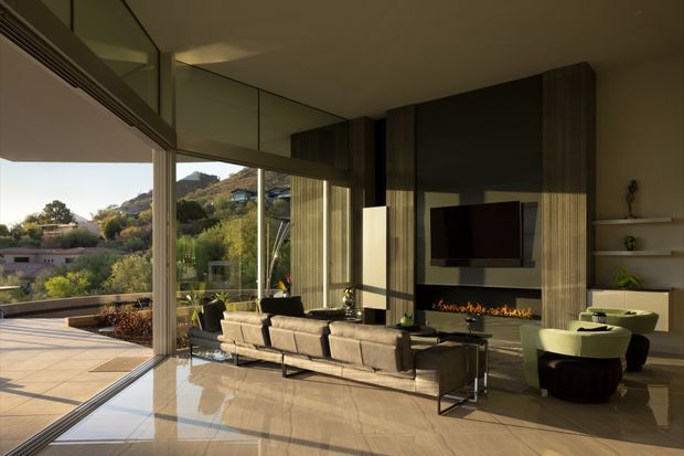 A sophisticated contemporary living room with floor-to-ceiling glazing opening onto a spacious terrace and landscaped hillside views. The room features a large sectional sofa facing a wall-mounted television and linear fireplace, dark vertical timber or wood-look wall panelling, integrated shelving, polished light stone flooring, and sculptural lounge chairs. Abundant natural light and expansive glazing create a strong indoor-outdoor connection.