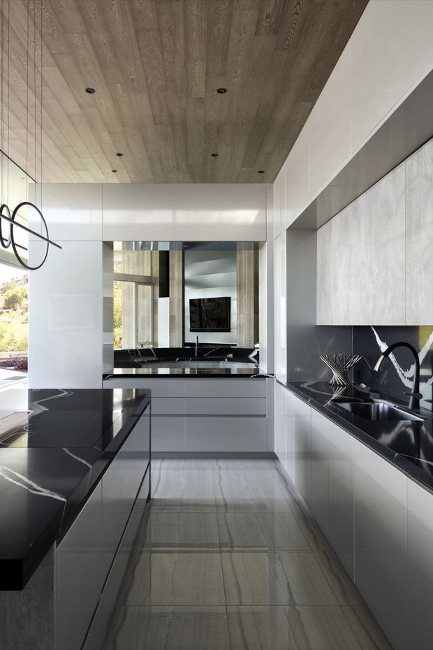 A sleek contemporary galley kitchen with white full-height cabinetry, glossy black stone benchtops and splashback, integrated sink and black tapware, and a large black stone island. The space features a warm timber-look recessed ceiling with downlights, polished light stone flooring, extensive reflective surfaces, and a mirror-like opening that visually connects to an adjacent living area.