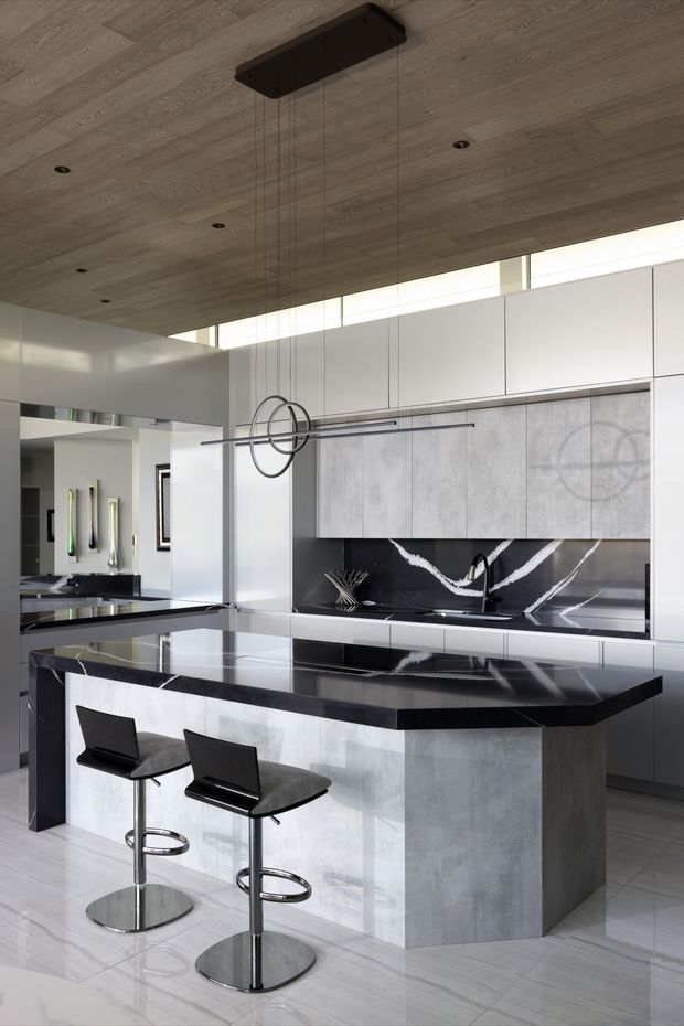 Contemporary kitchen dominated by a large central island with a dark polished stone benchtop, pale concrete-look cabinetry, and a dramatic black-and-white veined splashback. The room has glossy light flooring, extensive white overhead cabinetry, a timber-lined ceiling with recessed downlights, clerestory glazing, and sculptural linear pendant lighting. Two black bar stools sit at the island.