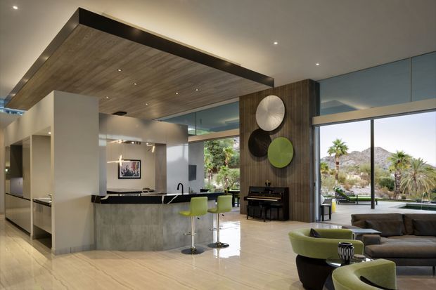 Open-plan contemporary living area with an integrated kitchen and breakfast island, featuring a dramatic suspended timber-lined ceiling, pale stone or concrete-look surfaces, dark timber wall panelling, floor-to-ceiling glazing, and views to a landscaped exterior. A piano and sculptural circular wall decor sit beside the living zone, which includes a dark sectional sofa and rounded lime-green accent chairs.