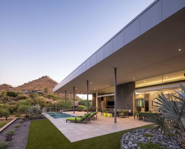 Exterior view of a low-slung contemporary residence with a broad flat roof and deep covered terrace. Floor-to-ceiling glazing opens onto a paved outdoor living area with lounge seating, a narrow swimming pool, artificial lawn, gravel landscaping, and sculptural desert plants. Rugged low hills are visible in the background.