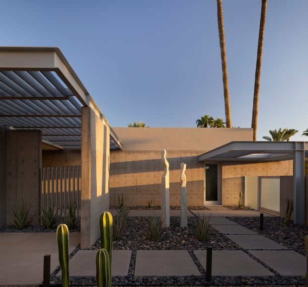 A low-slung modern courtyard exterior in warm concrete tones, featuring a flat-roofed building, exposed concrete structural posts, a slatted metal pergola and carport-like canopy, paved stepping paths, and desert xeriscape planting with cacti and agaves. Tall palm trees rise behind the walls against a clear blue sky.