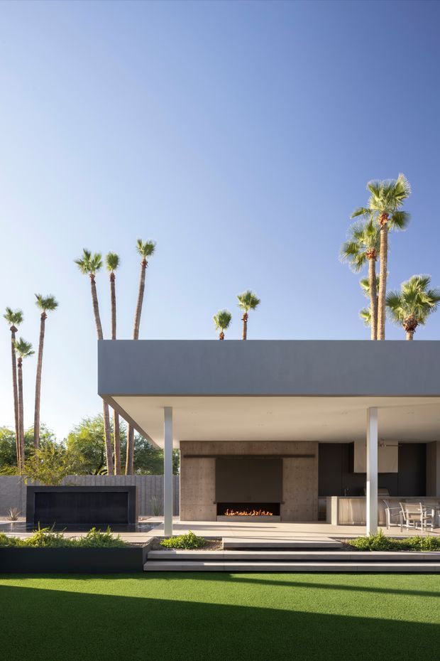 A contemporary flat-roofed outdoor entertaining area with a broad covered terrace, slim square columns, an open kitchen and dining space, and a built-in linear fireplace set into a large concrete-toned feature wall. Stepped paving leads to a manicured lawn, with tall palm trees surrounding the property. The composition is bright and minimal, with strong horizontal rooflines and indoor-outdoor living.