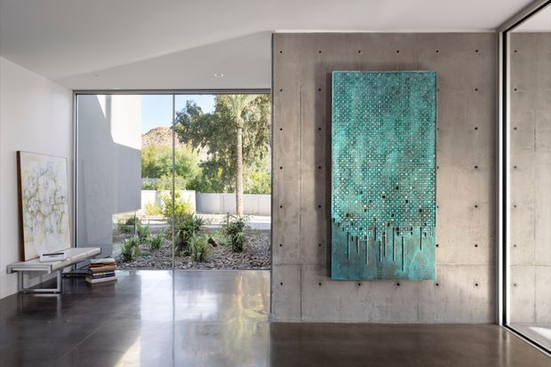 Contemporary minimalist interior with a polished dark concrete floor, smooth concrete feature wall, and full-height glazing opening onto a landscaped courtyard. A turquoise abstract wall artwork provides the main accent, while a low console, books, and framed artwork occupy the adjoining living area.